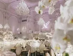 The image depicts the luxurious interior of Maison Principale, a wedding venue featured in the Mariage Elegant directory in Canada. The venue showcases an opulent setting with grand chandeliers hanging from a high, ornately decorated ceiling. Elegant floral arrangements add a touch of romance, complementing the classic round tables and plush seating, which are perfectly set up for a sophisticated wedding reception. The lighting casts a soft, ambient glow, highlighting the venue's exquisite architectural details, making it an ideal choice for couples seeking a memorable wedding venue.