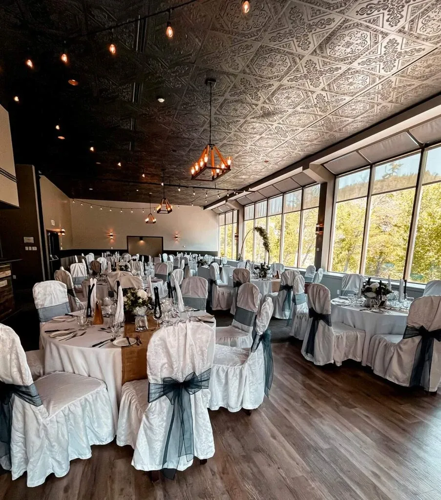 The image shows an elegantly decorated wedding venue at The Auberge Val Saint-Côme, a vendor in the Wedding Venues category. The spacious room features a beautifully embossed dark ceiling and large windows that let in natural light. Round tables are dressed in white tablecloths with intricate centerpieces, and each chair is covered with a white slipcover tied with a dark gray sash. Overhead, warm pendant lights contribute to the inviting atmosphere, making it an ideal setting for a wedding celebration.