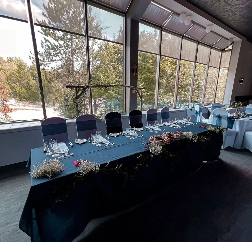 In this image, a beautifully set table is located in a charming wedding venue at The Auberge Val Saint-Côme, categorized under Wedding Venues. The table is elegantly draped with a dark tablecloth and adorned with delicate floral arrangements featuring baby's breath and greenery, creating a romantic ambiance. Each place setting includes neatly folded napkins and polished dinnerware. Large windows in the background flood the room with natural light and provide a scenic view of lush evergreen trees, enhancing the venue's serene and picturesque atmosphere. The chairs are covered with sleek white covers and blue sashes, adding a sophisticated touch to the overall decor.