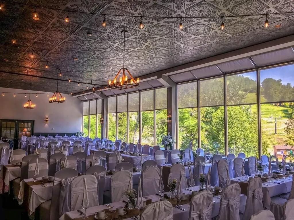 The image showcases the interior of The Auberge Val Saint-Côme, a wedding venue, set up for a wedding reception. The venue features rows of elegantly arranged banquet tables adorned with white tablecloths and intricate chair covers. Each table is decorated with delicate flower arrangements and fine china, creating a sophisticated and inviting atmosphere. The room is illuminated by unique, stylish chandeliers and has large windows allowing natural light to flood in, offering a picturesque view of the lush greenery outside. The ceiling has an ornate design, adding to the luxurious ambiance perfect for a wedding celebration at this Canadian venue.