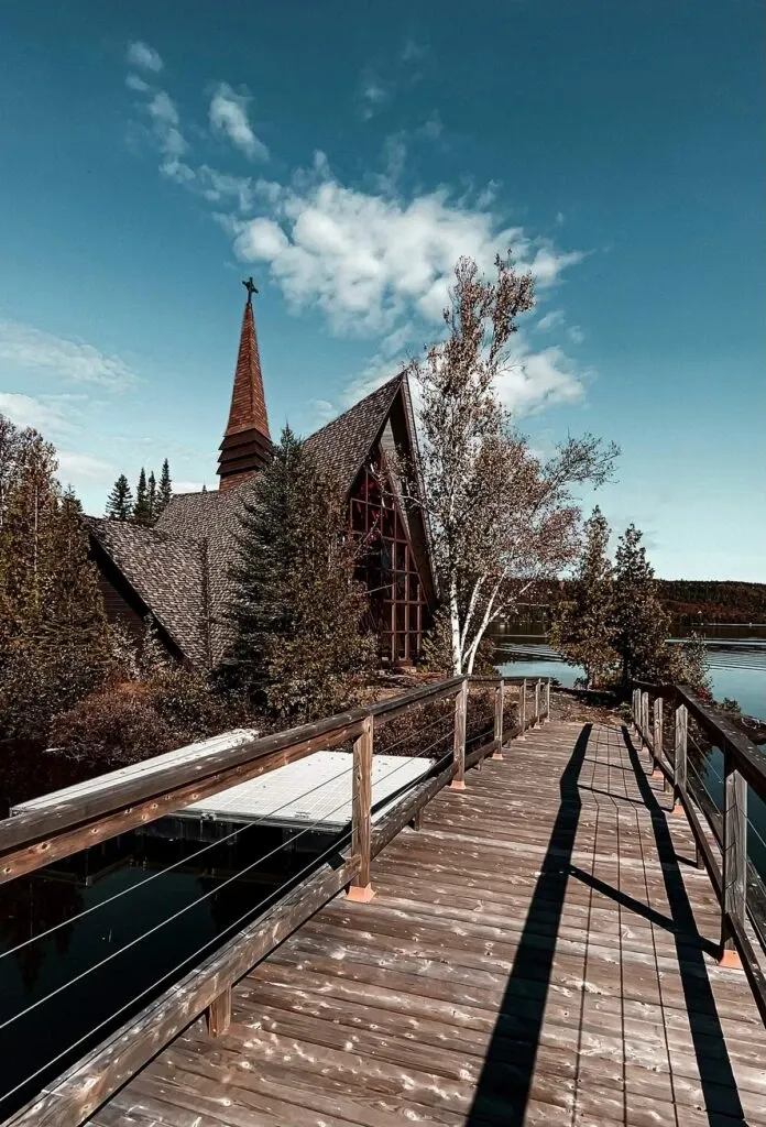 The image showcases The Auberge Val Saint-Côme, a charming wedding venue, surrounded by lush trees and a serene lake in Canada. The picture captures a rustic wooden walkway leading towards an elegant, chapel-like building with a steeply pitched roof and a tall spire. The structure is nestled amidst a setting of natural beauty, with a bright blue sky and fluffy clouds overhead. This idyllic scene highlights the venue's picturesque and tranquil atmosphere, perfect for a romantic wedding celebration.