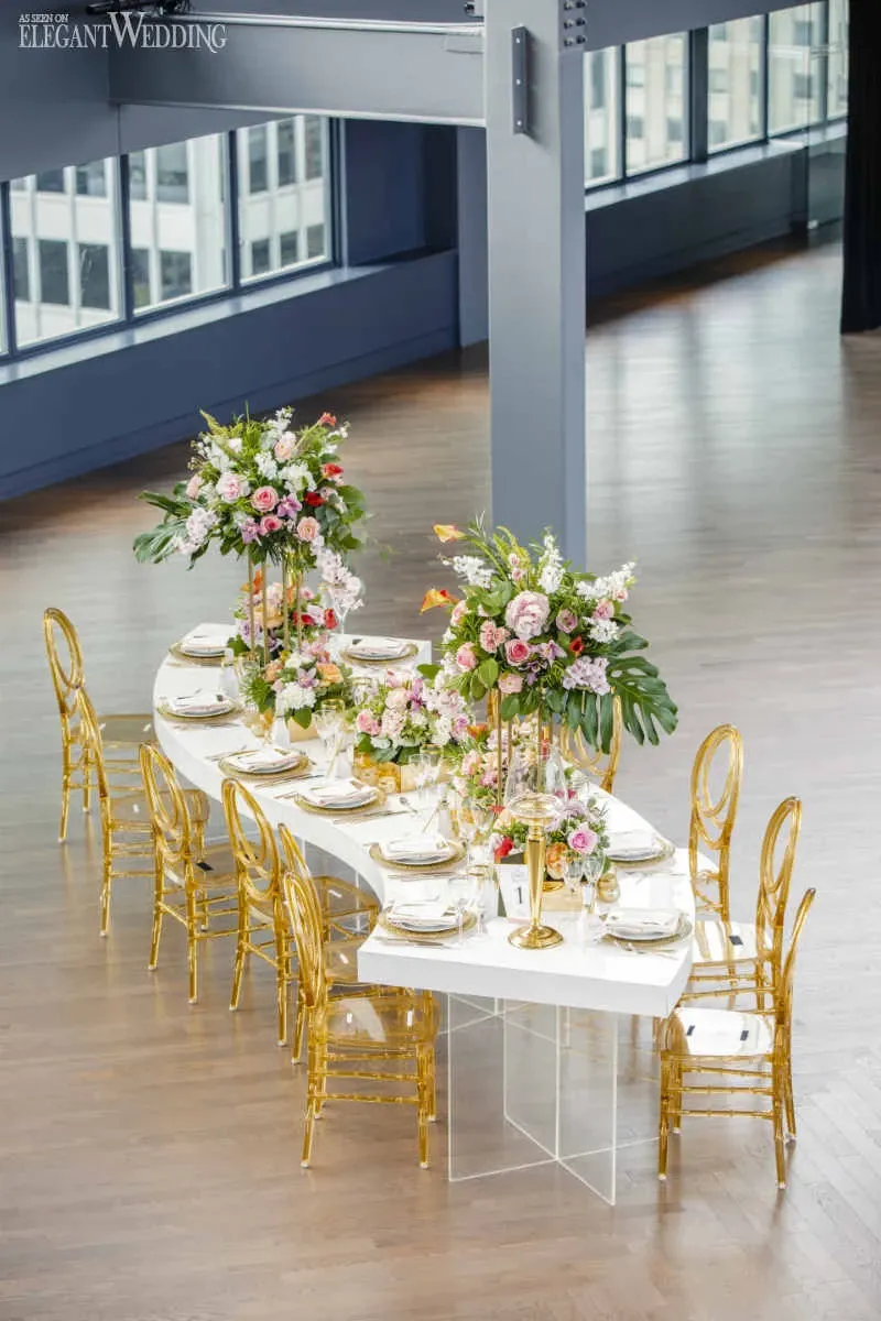 The image features a beautifully set wedding venue at Fairmont The Queen Elizabeth, categorized under Wedding Venues on Mariage Elegant. A sophisticated, elongated white table is elegantly arranged with luxurious floral centerpieces comprising pink, white, and green foliage. Surrounding the table are opulent, transparent gold chairs, complementing the modern and chic interior of the venue. Large windows in the background flood the space with natural light, highlighting the polished wooden floor and creating an airy, inviting atmosphere perfect for a wedding celebration.