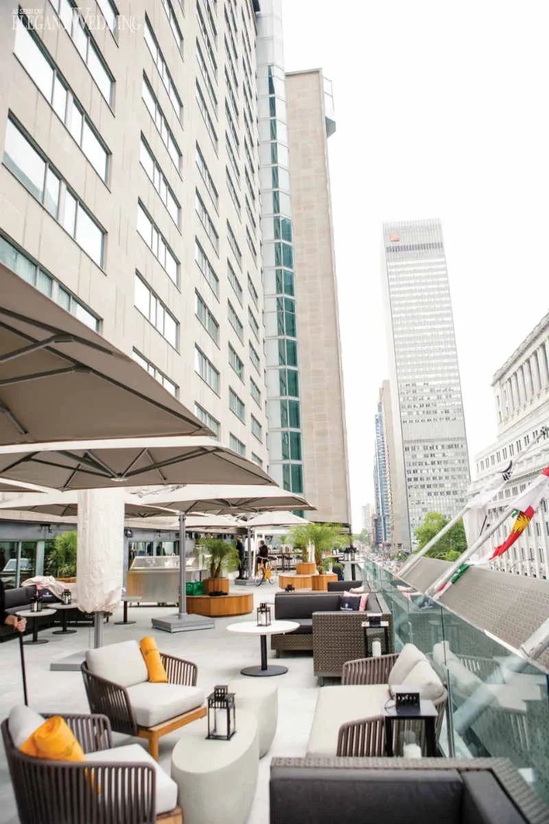 The image showcases the outdoor terrace of Fairmont The Queen Elizabeth, a prominent wedding venue in Canada. The terrace features elegant seating arrangements with modern sofas and plush throw pillows in shades of white, brown, and mustard. Large umbrellas provide shade, creating a refined and inviting atmosphere for guests. Tall buildings are visible in the background, adding an urban backdrop to this sophisticated wedding setting. The terrace's layout and decor make it an ideal spot for hosting wedding receptions or cocktail hours.