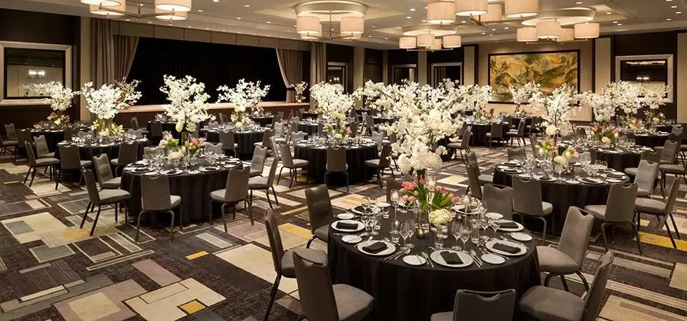 The image showcases a beautifully decorated wedding venue at Fairmont The Queen Elizabeth, categorized under Wedding Venues. The venue features elegantly arranged round tables covered with black tablecloths and set with fine china and glassware. Each table is adorned with tall, stunning floral centerpieces composed of white blossoms, adding a touch of elegance and sophistication to the room. The spacious room is well-lit with modern chandeliers and features a stage at the front, suitable for wedding ceremonies or receptions. The floor is designed with a colorful, geometric patterned carpet that complements the overall luxurious ambiance.