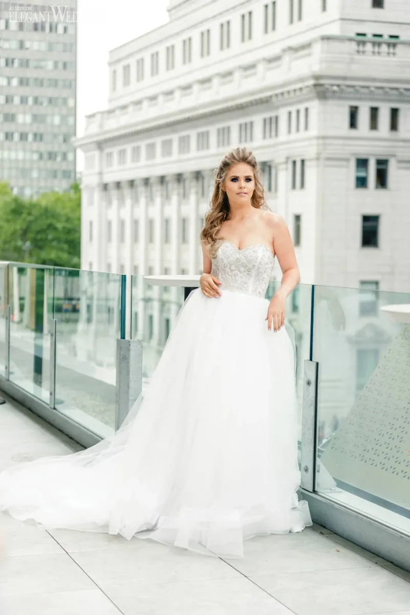 A bride stands elegantly on the rooftop terrace of Fairmont The Queen Elizabeth, a premium wedding venue in Canada. She is wearing a strapless, embellished white wedding gown that flows beautifully to the floor. The terrace features a glass railing, offering a stunning backdrop of a historic, grand building and lush greenery in the distance, suggesting a perfect urban setting for a sophisticated wedding ceremony or reception at this prestigious venue.