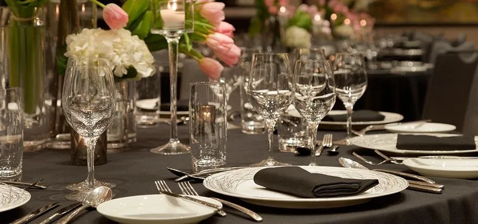 The image showcases an elegantly set table at Fairmont The Queen Elizabeth, categorized under wedding venues. The table features a sophisticated arrangement with black napkins neatly folded atop white china plates, complemented by an array of glassware including wine and water glasses. Silver cutlery is meticulously placed, adding a touch of refinement. The centerpiece includes vibrant pink tulips and soft white hydrangeas placed in clear, refined vases, contributing to an ambiance of romance and luxury, ideal for a wedding celebration.