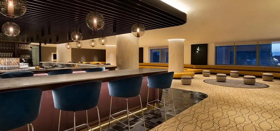 This image features an elegant and modern interior view of Fairmont The Queen Elizabeth, categorized as a wedding venue on Mariage Elegant, Canada. The venue showcases a stylish bar area with blue upholstered bar stools neatly aligned along a polished countertop. Above the bar, several round pendant lights emit a warm glow, creating a sophisticated ambiance. The room also includes plush seating areas with round ottomans and large windows offering a view of the cityscape at dusk. This setting provides a chic and versatile space, ideal for hosting wedding receptions or related events.