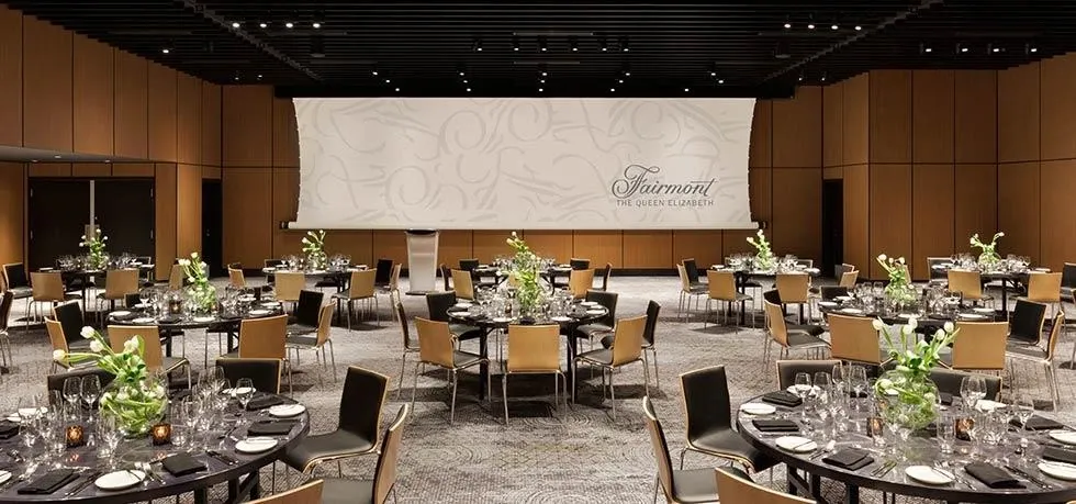 The image shows an elegant wedding venue at Fairmont The Queen Elizabeth, featuring a spacious banquet hall. Round tables are neatly arranged across the room, each adorned with white linens, tasteful green and white floral centerpieces, and set with polished glassware and cutlery. The ambiance is modern and sophisticated, with a neutral color palette. A large projection screen at the front displays the name 'Fairmont The Queen Elizabeth,' emphasizing the venue's prestigious brand. This venue provides a refined setting suitable for weddings, accommodating a significant number of guests comfortably.
