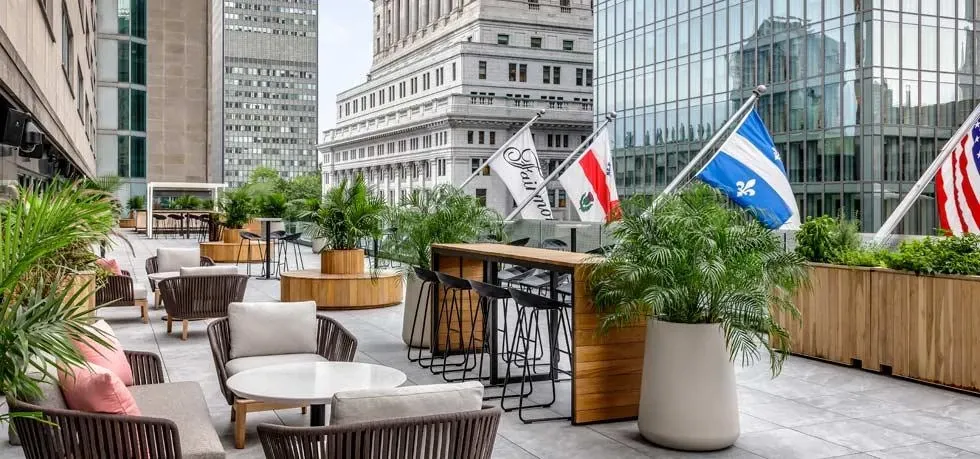 The image shows an elegant outdoor terrace at Fairmont The Queen Elizabeth, a wedding venue, featuring modern wicker chairs with soft cushions and round tables set against a backdrop of lush greenery. The terrace overlooks a cityscape, with several prominent office buildings visible. Three flags, including a Québécois flag, are prominently displayed, adding a touch of local flair. The setup provides a sophisticated ambiance, ideal for outdoor wedding receptions or ceremonies.