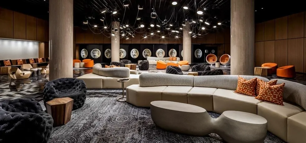 The image showcases a modern and stylish interior of Fairmont The Queen Elizabeth, a renowned wedding venue. The space features a contemporary lounge area with plush, dark velvet seating and unique, curved beige couches accentuated by orange patterned throw pillows. The floor displays a chic, geometric pattern that adds to the elegant ambiance. Overhead, sleek lighting fixtures hang from the ceiling, creating a warm and inviting atmosphere. In the background, circular wall art adds a decorative touch, while orange and neutral tones dominate the color palette, making it an ideal setting for a sophisticated wedding event.