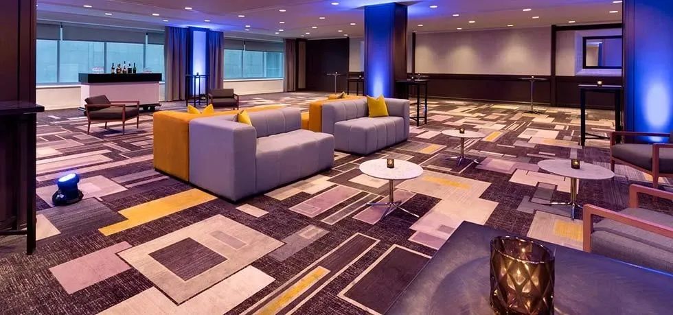 This image features an elegantly decorated indoor space at Fairmont The Queen Elizabeth, categorized under Wedding Venues on Mariage Elegant, Canada. The spacious room showcases modern, geometric-patterned carpeting in shades of gray, yellow, and beige, complementing the ambient lighting. Contemporary gray and yellow sofas are arranged alongside sleek, round coffee tables, creating an inviting seating area. The large windows in the background allow natural light to flow in, enhancing the warm and sophisticated ambiance, perfect for hosting intimate wedding events.