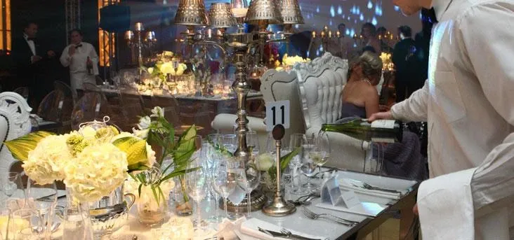 The image shows an elegantly set table at Fairmont The Queen Elizabeth, a wedding venue in Canada. The table is adorned with white floral arrangements and lush green leaves, surrounded by fine glassware and silver cutlery, signaling a sophisticated wedding setting. A server is seen pouring wine into glasses, while guests in formal attire mingle in the softly lit background. The number '11' is displayed on a stand, likely indicating the table number. This setting exemplifies the luxurious ambiance expected at a wedding hosted at this prestigious venue.