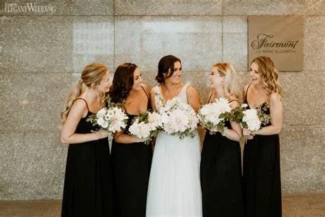 A bride stands with four bridesmaids in front of a granite wall at the Fairmont The Queen Elizabeth, under a sign displaying the venue's name. The bride is wearing a white gown and holds a bouquet of white and blush flowers, while the bridesmaids are dressed in black dresses, each holding similar bouquets. This elegant scene captures the sophisticated and timeless aesthetic of a wedding venue at Fairmont The Queen Elizabeth in Canada.