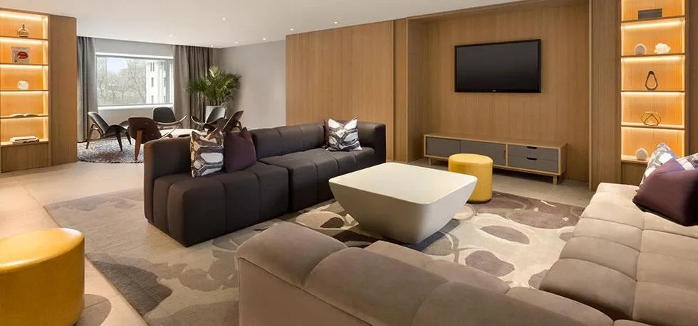The image showcases an elegantly designed lounge area inside Fairmont The Queen Elizabeth, a wedding venue featured on Mariage Elegant, Canada. The room is furnished with modern sofas in neutral tones arranged around a stylish, geometric coffee table. On the right wall, a large flat-screen TV is mounted above a sleek, low-standing cabinet. The walls are adorned with built-in shelves displaying decorative items softly lit with ambient lighting. A large window on the left lets in natural light, enhancing the modern and inviting atmosphere. This sophisticated setting serves as an ideal space for wedding gatherings and celebrations.