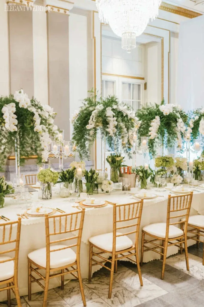 The image showcases a beautifully set wedding reception at Le Windsor Ballrooms, a premier wedding venue in the Mariage Elegant vendor listing in Canada. The scene features a long, elegantly decorated table adorned with lush white and green floral arrangements cascading from tall glass vases. Gold accents are prominent, with elegant gold-rimmed plates and matching gold cutlery. Delicate candle arrangements add a soft glow, enhancing the luxurious ambiance. Classic wooden Chiavari chairs with white cushions are neatly arranged around the table, complementing the refined aesthetic of the venue. The background reveals high ceilings with intricate moldings and a stunning crystal chandelier, contributing to the sophisticated atmosphere of this wedding setting.