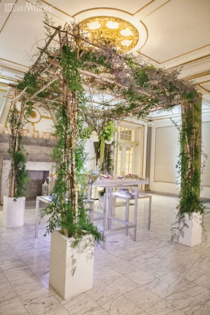 The image showcases a beautifully decorated interior space at Le Windsor Ballrooms, a premier wedding venue in Canada. The room features a stunning square arch structure adorned with lush green foliage and delicate branches, creating a natural, romantic ambiance. The arch is elegantly positioned over a white high-top table, enhancing the sophisticated setting perfect for a wedding ceremony or reception. The marble floor and ornate ceiling with a central gold chandelier add a touch of luxury and grandeur, reinforcing the venue's reputation as a top choice for elegant weddings.