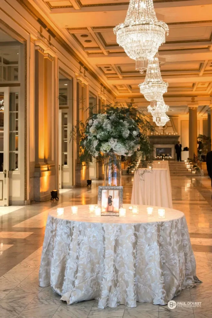 The image showcases an elegant interior of Le Windsor Ballrooms, a premier wedding venue in Canada, categorized under wedding venues on Mariage Elegant. The photograph captures a lavishly decorated ballroom featuring a round table draped with a detailed white tablecloth adorned with intricate floral patterns. On top of the table, there is a stunning floral arrangement in a tall vase, accompanied by a framed picture and surrounded by small, glowing tea light candles. The opulent setting is highlighted by grand chandeliers hanging from the ceiling, casting a warm and inviting light over the marble floors and ornate walls. The ambiance exudes sophistication and luxury, perfect for an unforgettable wedding celebration.