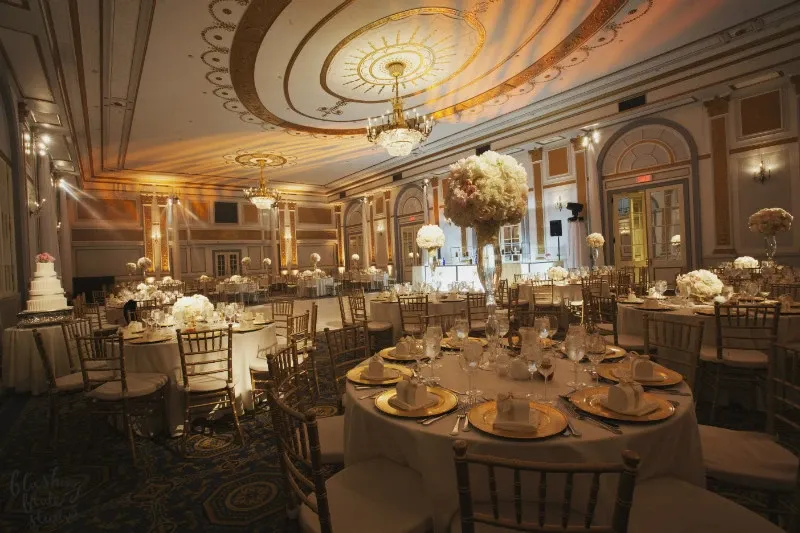 The image showcases the elegant Le Windsor Ballrooms, highlighting its grandeur as a premier wedding venue. The spacious ballroom is beautifully decorated with round tables draped in white linens, set with fine china, crystal glassware, and gold cutlery. Tall floral centerpieces with white and pink flowers add a romantic touch to each table. The room's opulent decor features intricate ceiling designs, soft ambient lighting, and large windows draped with luxurious curtains, creating a sophisticated and inviting atmosphere perfect for a wedding celebration.