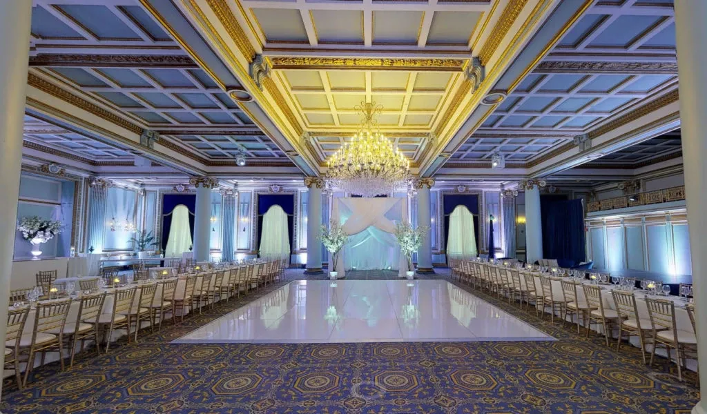 The image showcases Le Windsor Ballrooms, a distinguished wedding venue in Canada, under the Mariage Elegant platform. The ballroom is elegantly set up for a wedding, featuring a polished white dance floor at the center, surrounded by opulent gold-trimmed columns. Rows of lavishly decorated tables with gold Chiavari chairs line each side of the dance floor, ready for guests. Above, an exquisite crystal chandelier hangs from the ceiling, casting a warm glow and enhancing the grandeur of the ornately coffered ceiling. Floral arrangements and soft blue uplighting create a romantic atmosphere, ideal for a wedding celebration.