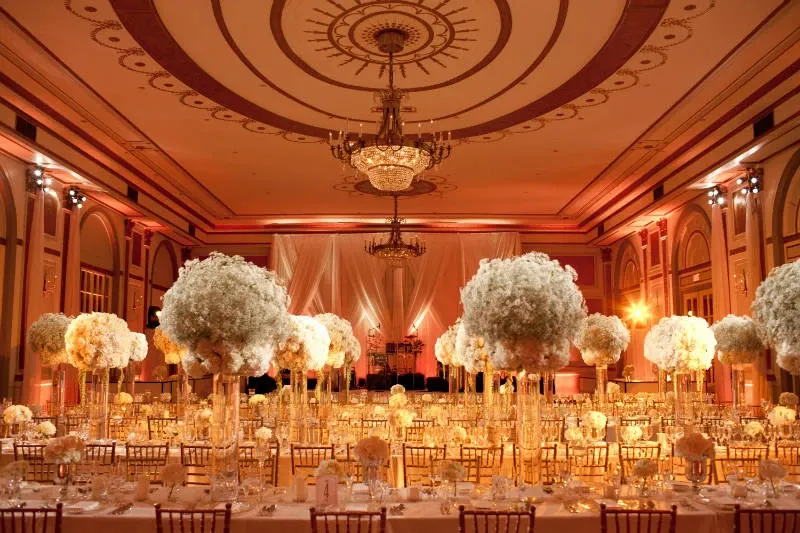 The image showcases the opulent interior of Le Windsor Ballrooms, a prominent wedding venue in Canada. The spacious ballroom is elegantly adorned with grand chandeliers hanging from a richly decorated ceiling. Rows of tables are set for a wedding reception, featuring stunning floral centerpieces with tall, white flower arrangements. The warm lighting creates a luxurious and inviting atmosphere, highlighting the grandeur of the venue.
