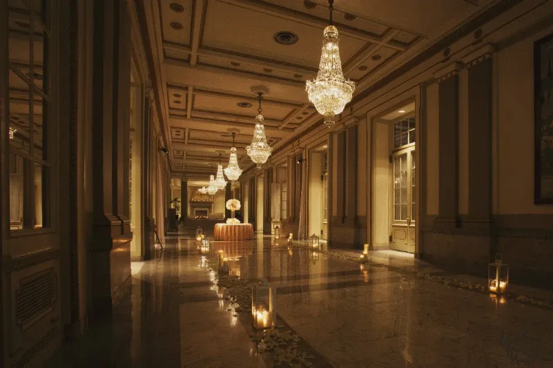 This image showcases the elegant interior of Le Windsor Ballrooms, a premier wedding venue in Canada. The grand hallway is adorned with exquisite crystal chandeliers that cast a warm, inviting glow over the polished marble floors. The space is beautifully decorated with numerous candles lining the pathway and leading to a sophisticated flower arrangement centerpiece on a table. The classic architectural details, including ornate molding and tall windows, enhance the luxurious ambiance, making it a perfect setting for a memorable wedding celebration.