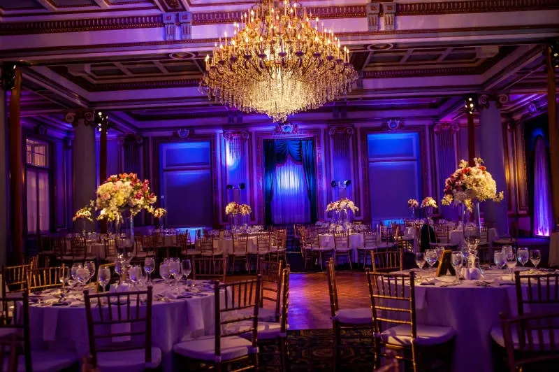 The image depicts a luxurious wedding venue at Le Windsor Ballrooms, belonging to the Wedding Venues category, featured on Mariage Elegant, Canada. The venue showcases an opulent ballroom setting adorned with an extravagant crystal chandelier hanging from an intricately designed ceiling. Elegant round tables are set with fine china, glassware, and tall, floral centerpieces, adding a touch of sophistication. The warm, ambient lighting and royal purple uplighting create a romantic and grand atmosphere, perfect for an upscale wedding celebration. Gold Chiavari chairs encircle the tables, completing the elegant décor.