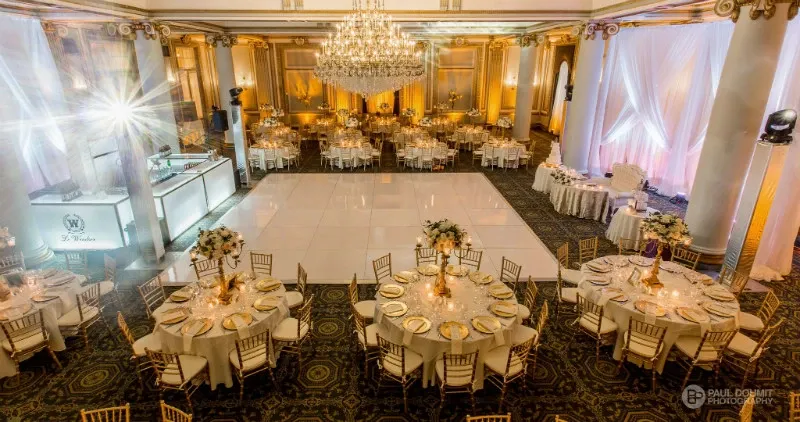 The image showcases the elegant interior of Le Windsor Ballrooms, a premier wedding venue in Canada, featured on Mariage Elegant. The spacious ballroom is adorned with large, opulent chandeliers casting a warm glow over immaculately set round tables, each draped with white linens and surrounded by gold Chiavari chairs. Floral centerpieces and delicate table settings enhance the sophisticated atmosphere, perfect for a wedding reception. A polished dance floor occupies the center of the room, inviting guests to celebrate in style. Tall columns and subtly lit white drapery along the walls add to the grandeur of the venue.