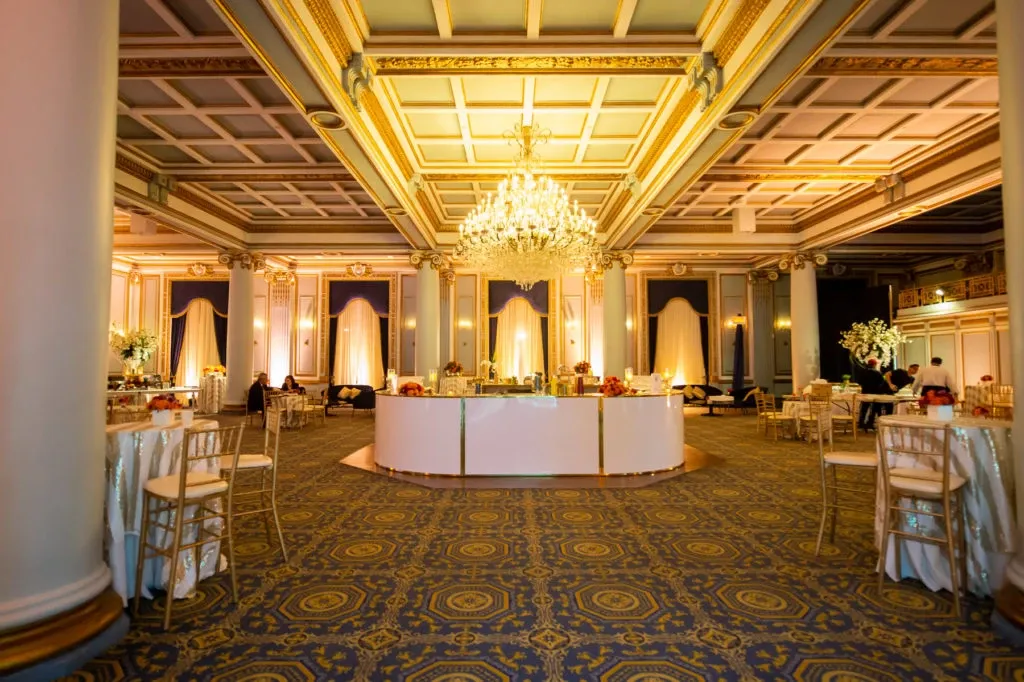 The image showcases the opulent interior of Le Windsor Ballrooms, a prestigious wedding venue listed on Mariage Elegant, Canada. The grand ballroom features an exquisite chandelier hanging from a beautifully coffered ceiling, casting a warm glow over the room. Elegant gold and white adornments accentuate the space, with tall pillars and detailed moldings enhancing its classic charm. Round tables are elegantly set with white tablecloths, gold chiavari chairs, and subtle floral centerpieces, creating a sophisticated atmosphere ideal for a wedding celebration. The luxurious decor and spacious layout make Le Windsor Ballrooms a perfect choice for hosting memorable weddings.