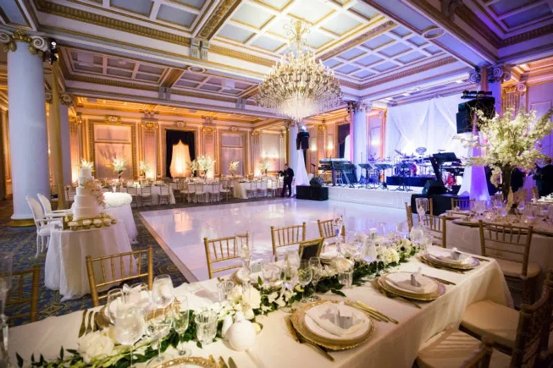 The image showcases a luxurious wedding venue at Le Windsor Ballrooms, categorized as a wedding venue on Mariage Elegant, Canada. The scene is set with an elegant ballroom featuring a grand chandelier hanging from an intricately designed ceiling. A spacious, shiny white dance floor is centered, surrounded by elegantly set tables adorned with white and gold tableware, floral centerpieces, and soft candlelight. The room is illuminated with warm, ambient lighting, highlighting the gold detailing on the columns and the opulent decor. On one side, a stage is set up with musical instruments, ready for live performances, creating a perfect setting for a sophisticated wedding reception.