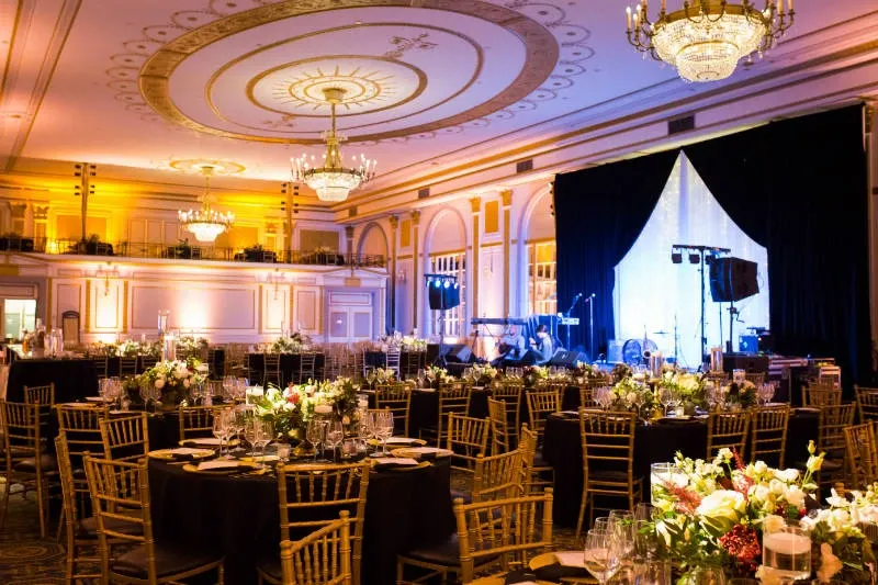 The image showcases the elegant Le Windsor Ballrooms, categorized under wedding venues for Mariage Elegant, Canada. The ballroom is beautifully adorned with ornate chandeliers and intricate ceiling designs. The tables are set with fine china and glassware, surrounded by golden chiavari chairs. Each table is elegantly decorated with lush floral centerpieces. The warm lighting creates a romantic and inviting ambiance, perfect for a sophisticated wedding reception. A stage is visible in the background, prepared for entertainment or speeches, adding to the venue's readiness for a memorable event.