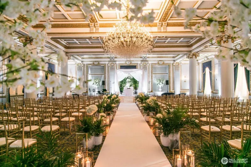 The image features the elegant Le Windsor Ballrooms, a wedding venue situated in Canada, showcasing a grand hall beautifully decorated for a wedding ceremony. The aisle is lined with lush greenery and white flowers, leading to an intricately carved white altar with decorative drapery. The ceiling boasts opulent gold and white detailing, complemented by a stunning crystal chandelier that illuminates the room. Rows of gold chiavari chairs are arranged neatly, offering ample seating for guests. The setting exudes sophistication, perfect for a luxurious wedding celebration.