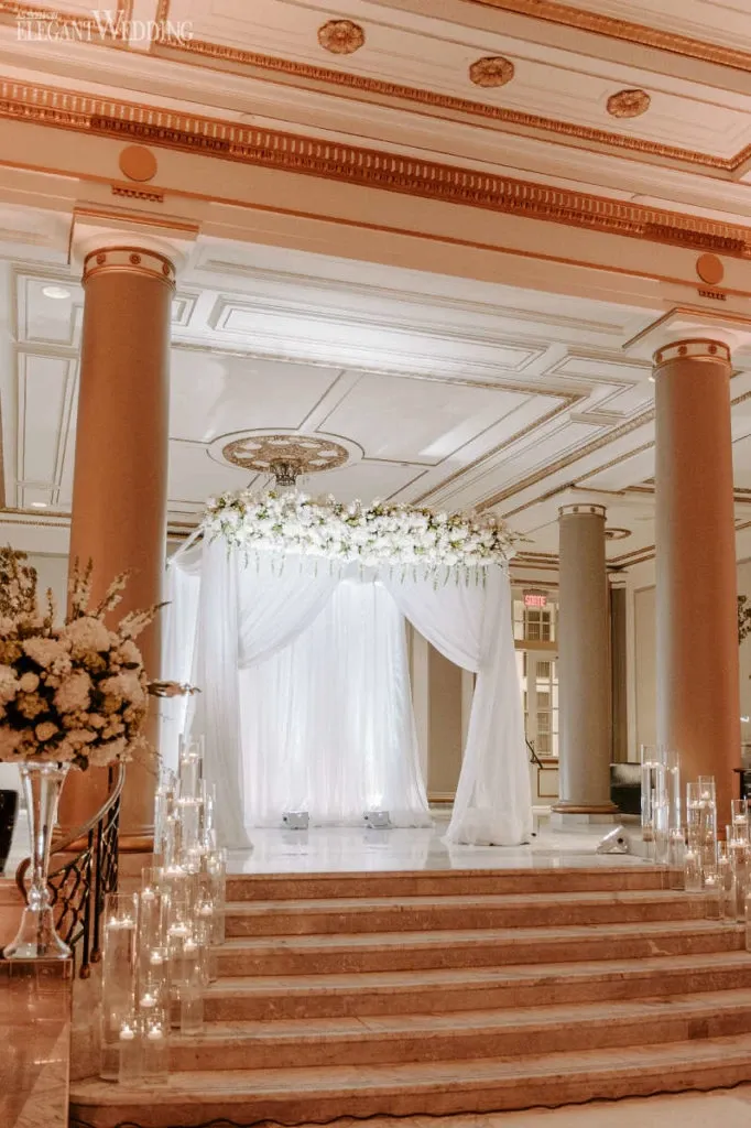 The image showcases a beautifully decorated wedding venue at Le Windsor Ballrooms, a premier wedding venue in the wedding planning and vendor listing category for Mariage Elegant, Canada. The setting features an elegant altar adorned with flowing white drapery and floral arrangements. The altar is situated on a raised platform with marble steps, surrounded by tall, illuminated candles in glass holders. The venue's classic architectural elements, including ornate ceiling moldings and grand columns, add a sophisticated ambiance, making it an ideal choice for weddings.