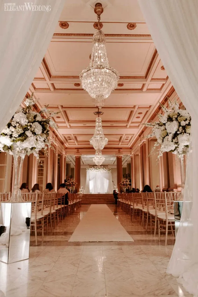 This image showcases the elegant interior of Le Windsor Ballrooms, a premier wedding venue in Canada. The venue features a grand aisle lined with luxurious white fabric and bordered by tall, exquisite floral arrangements in shades of white and cream, adding a touch of sophistication. The scene is enhanced by the opulent crystal chandeliers hanging from the ceiling, casting a warm, inviting glow over the guests seated on either side in gold chairs. The ambiance is perfect for a wedding ceremony, capturing the timeless elegance and grandeur that Le Windsor Ballrooms offers for couples planning their special day.