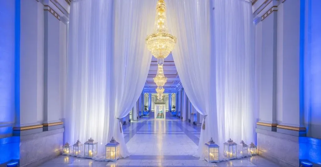 The image showcases the Le Windsor Ballrooms, a premier wedding venue in Canada, under the category of Wedding Venues on Mariage Elegant's site. The elegant ballroom features a grand entrance adorned with luxurious white drapes creating a sophisticated aisle. Extravagant chandeliers hang from the ceiling, illuminating the space with a warm, inviting glow. Alongside the aisle are numerous decorative lanterns, adding a romantic touch to the setting. The marble flooring complements the refined ambiance, making it an ideal location for upscale wedding ceremonies or receptions.