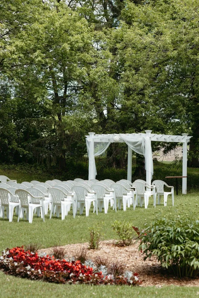 The image depicts a charming outdoor wedding setup at Manoir Grant, a renowned wedding venue in Canada. Rows of white chairs are neatly arranged facing a white pergola adorned with delicate, flowing white fabric, creating an elegant aisle for ceremonies. The setting is surrounded by lush green trees, providing a serene and natural backdrop. In the foreground, a beautifully maintained flower bed with vibrant red and white flowers adds a touch of color and charm to the venue, enhancing its appeal for wedding celebrations.