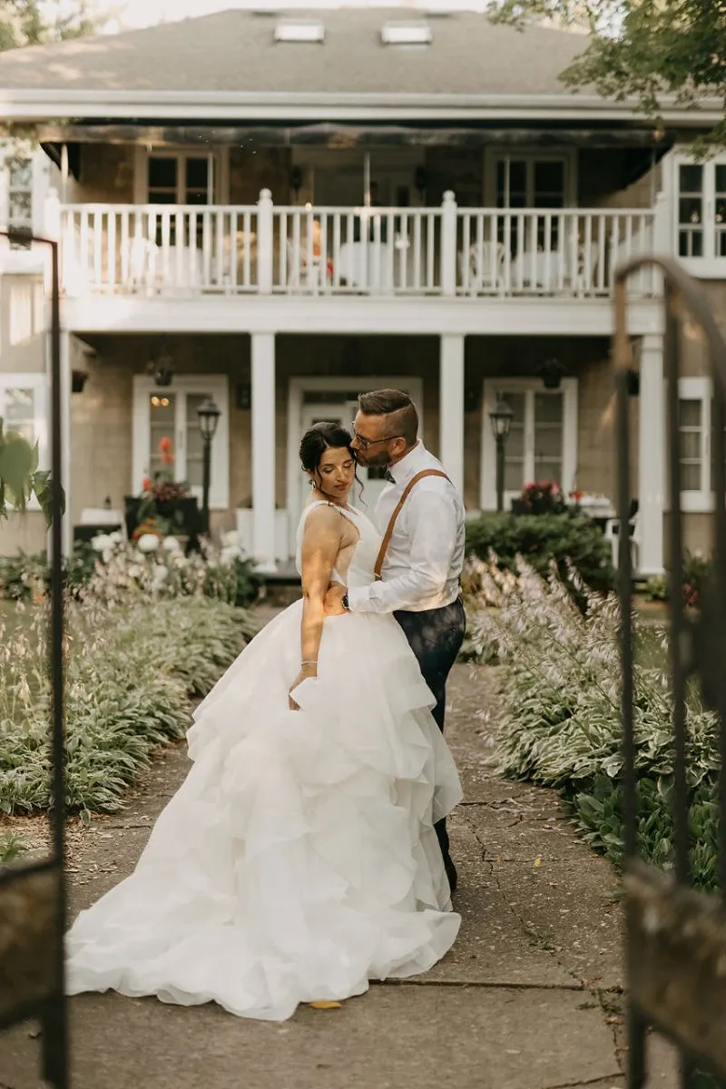 A bride and groom are standing in a garden path in front of the elegant Manoir Grant, a premier wedding venue in Canada. The bride is wearing a luxurious, layered white gown and the groom is dressed in formal attire with suspenders. They are embracing romantically with the backdrop of a charming, two-story building featuring a balcony adorned with flowers. The lush greenery and well-maintained garden enhance the romantic and picturesque setting, ideal for weddings and special events.