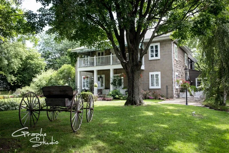 This image features the Manoir Grant, a charming wedding venue located in Canada, as part of the Mariage Elegant vendor listing. The scene depicts a picturesque, historic manor surrounded by lush greenery and mature trees, creating an idyllic setting for weddings. A vintage wooden carriage is placed on the well-manicured lawn, adding a touch of rustic elegance to the venue’s ambiance. The two-story stone building showcases large windows and a wrap-around balcony adorned with decorative railings, contributing to its classic appeal.