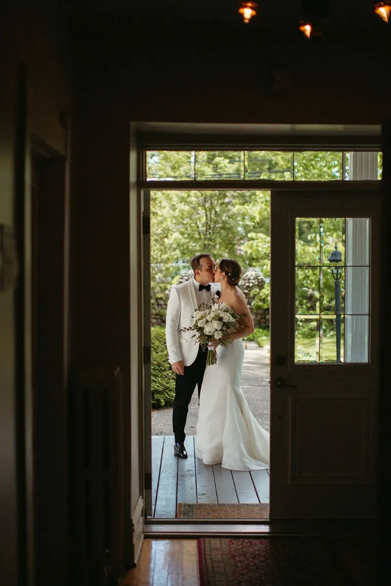 In this image, a newlywed couple shares a romantic kiss at the entrance of Manoir Grant, a featured wedding venue on Mariage Elegant, Canada. The groom, dressed in a white tuxedo jacket, embraces the bride, who wears an elegant white wedding gown and holds a lush bouquet of white and green flowers. They stand framed by a doorway that opens to a picturesque outdoor garden area, where greenery and soft lighting suggest an intimate, serene atmosphere ideal for weddings.
