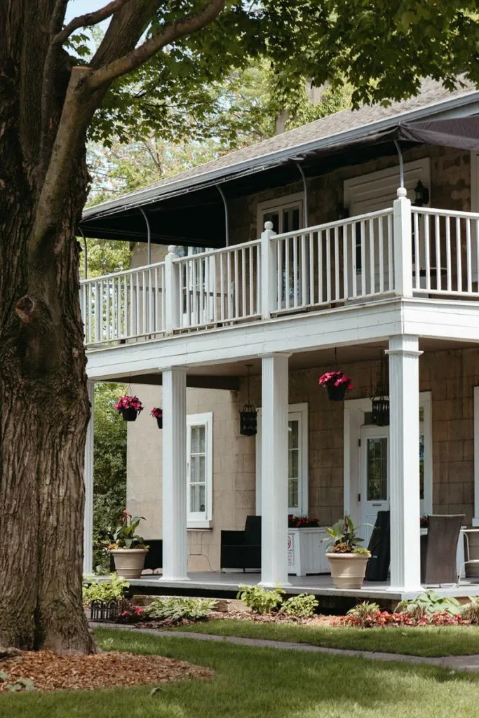 The image depicts Manoir Grant, a charming wedding venue in Canada, featuring a two-story building with a picturesque patio. The exterior showcases a rustic design with stone walls and a welcoming covered porch. Beautiful hanging planters with vibrant flowers adorn the porch roof, adding a touch of elegance. The lush greenery and blooming flower beds in the foreground create a serene and inviting setting, ideal for a romantic wedding celebration.