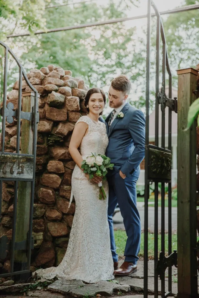 A bride and groom stand happily together in an outdoor setting at Manoir Grant, a renowned wedding venue in Canada. The bride, dressed in a detailed lace gown, holds a bouquet of white roses. The groom, wearing a sharp blue suit, lovingly looks at her. They are framed by an elegant wrought iron gate and rustic stone wall, reflecting the romantic and charming ambiance offered by this wedding venue. Sunlight filters through the trees, adding a soft, natural glow to the scene, perfect for a memorable wedding day.