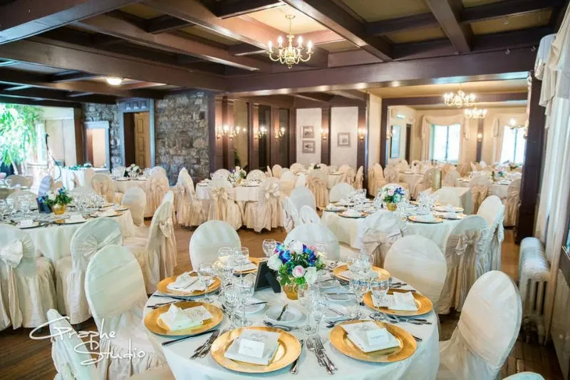 This image showcases the elegant wedding venue, Manoir Grant, categorized under wedding venues for Mariage Elegant in Canada. The reception area is beautifully arranged with round tables covered in pristine white tablecloths, each adorned with gold chargers and neatly folded napkins. Centerpieces featuring vibrant flowers enhance the tables, adding a touch of color and sophistication. The room's rustic charm is accentuated by stone walls and wooden beams on the ceiling, illuminated by classic chandeliers, creating a warm and inviting ambiance perfect for a wedding celebration.