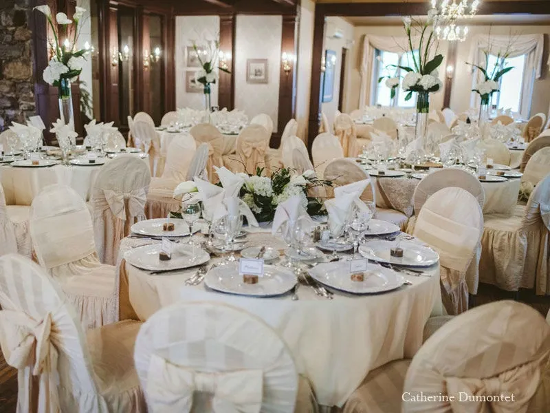 This image showcases an elegantly decorated wedding venue at Manoir Grant, a prestigious location in the Wedding Venues category on Mariage Elegant, Canada. The room features multiple round tables, each adorned with white tablecloths and neatly folded napkins atop fine china plates. The chairs are covered with matching cream-colored fabric, tied with large bows on the back. Arrangements of white flowers and lush greenery serve as table centerpieces, adding a touch of sophistication. The space is warmly lit by chandeliers and wall sconces, creating an inviting and romantic atmosphere, ideal for memorable celebrations.