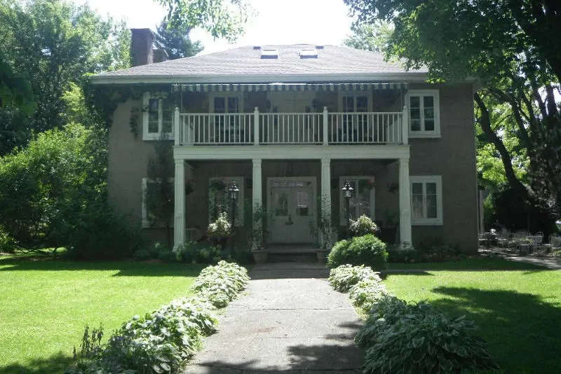 The image shows a charming view of Manoir Grant, a wedding venue in Canada, surrounded by lush greenery. The two-story building has a classic colonial style with a symmetrical façade and a welcoming covered porch adorned with elegant hanging lanterns. Beautifully manicured lawns and neatly trimmed shrubbery line the stone pathway leading towards the entrance, creating an inviting and serene setting ideal for wedding ceremonies and receptions. The backdrop of tall, mature trees adds to the intimate and picturesque atmosphere of this venue within the wedding planning category.