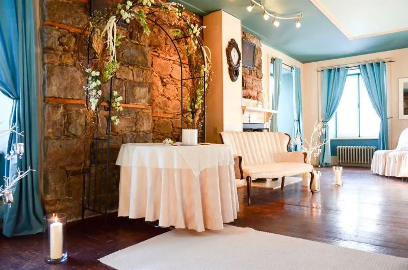 The image showcases a beautifully decorated wedding venue at Manoir Grant, a featured vendor under the Wedding Venues category on Mariage Elegant, Canada. The setting includes a charming indoor space with a rustic stone wall adorned with an elegant floral arch. A round table covered with a soft, white tablecloth sits in the foreground, surrounded by glowing pillar candles. In the background, a classic cream-colored loveseat is positioned next to a fireplace, adding a cozy ambiance. The room is tastefully accented with blue drapes and soft lighting, creating a romantic atmosphere ideal for a wedding celebration.