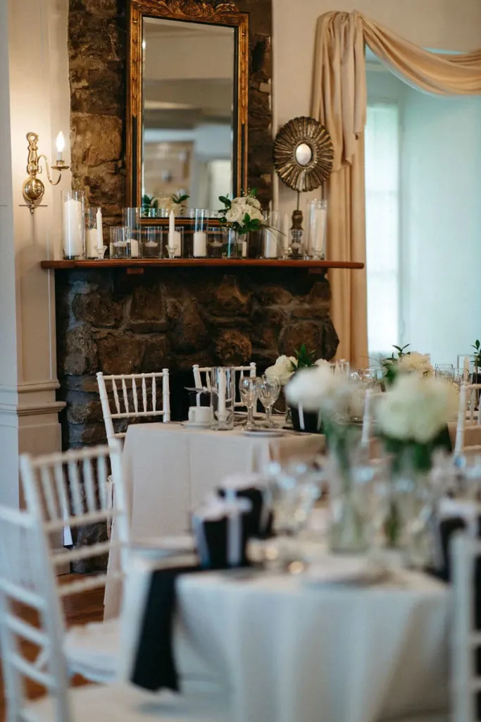 The image features an elegant indoor setting at Manoir Grant, categorized under Wedding Venues on Mariage Elegant, Canada. The room is adorned with white chiavari chairs surrounding tables draped in white linens, creating a sophisticated ambiance. The tables are elegantly set with glassware and floral centerpieces featuring white roses. A stone fireplace in the background is topped with white pillar candles and a mirror, adding to the venue's charm. Luxurious drapes hang at the windows, allowing soft natural light to add warmth to the setting, ideal for an intimate wedding celebration.