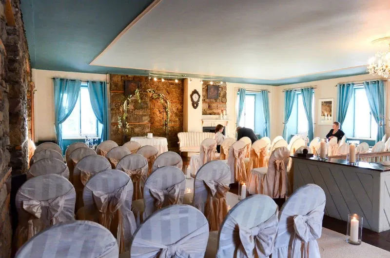 This image showcases an elegant wedding venue at Manoir Grant, categorized under Wedding Venues on Mariage Elegant, Canada. The room is beautifully decorated, featuring rows of white chairs with satin covers and intricate bow details, arranged for a wedding ceremony or reception. The soft blue curtains complement the ambient lighting, creating a serene atmosphere, while the stone wall adds a rustic charm. Elegant wall art and a cozy seating area with a white sofa enhance the venue's sophisticated look, ideal for a memorable wedding event.