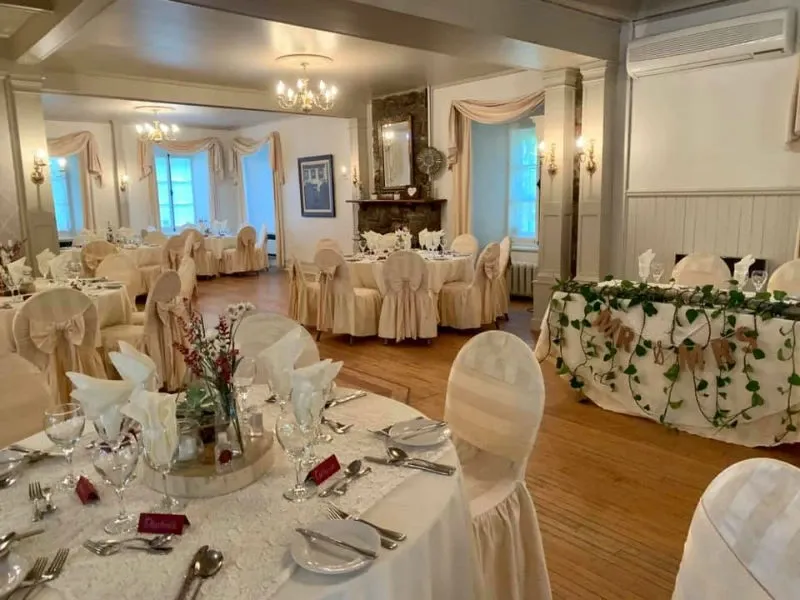 The image depicts a beautifully decorated wedding venue at Manoir Grant, categorized under Wedding Venues on the Mariage Elegant, Canada site. The elegantly arranged dining area features round tables adorned with white tablecloths, surrounded by chairs covered in beige fabric with decorative bows. Each table is set with pristine white napkins, glassware, and small floral arrangements, exuding a sophisticated ambiance. The room, illuminated by chandeliers, has soft white walls and delicate drapery, creating a warm and inviting atmosphere perfect for a wedding celebration. In the background, a long table is decorated with lush greenery, adding a touch of natural elegance to the setting.
