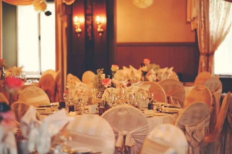 The image showcases the elegant interior of Manoir Grant, a wedding venue in the category of Wedding Venues listed on Mariage Elegant, Canada. The room is beautifully decorated with round tables draped in cream-colored tablecloths and chairs adorned with matching fabric covers and bows. Each table is set with fine glassware and adorned with delicate floral centerpieces featuring roses and baby's breath. The warm lighting from wall sconces creates an inviting and romantic atmosphere, while sheer curtains softly filter natural light from the windows. This setting exemplifies a perfect venue for an intimate and sophisticated wedding celebration.