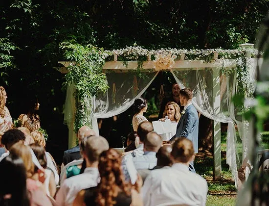 The image shows an outdoor wedding ceremony at Manoir Grant, a picturesque wedding venue in Canada. A couple stands under a beautifully decorated white pergola adorned with lush greenery and delicate floral arrangements, exchanging vows in front of a seated audience. The sunlight filters through the foliage, casting dappled shadows on the guests, who are attentively watching the ceremony. The setting is tranquil and elegant, embodying the romantic atmosphere that Manoir Grant offers for wedding celebrations.