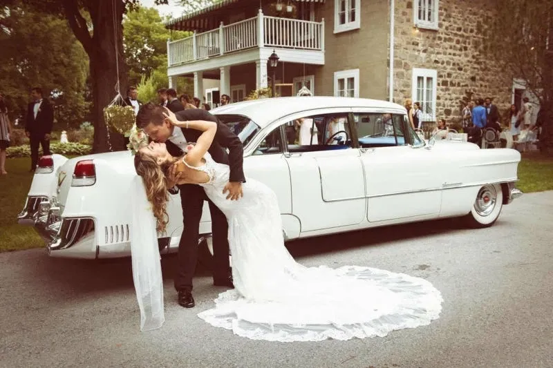 A bride in a flowing white gown and a groom in a classic black suit share a romantic kiss in front of a vintage white car, parked outside an elegant stone building with a balcony. This picturesque scene takes place at the Manoir Grant, a charming wedding venue located in Canada, ideal for couples seeking a blend of historical charm and scenic beauty for their special day. The lush greenery and well-dressed guests in the background add to the celebratory atmosphere, highlighting the venue's suitability for memorable wedding events.